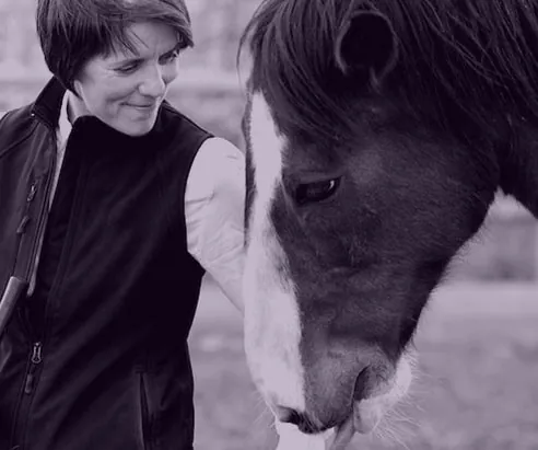 A woman smiling at a grey horse, in a purple-toned photo